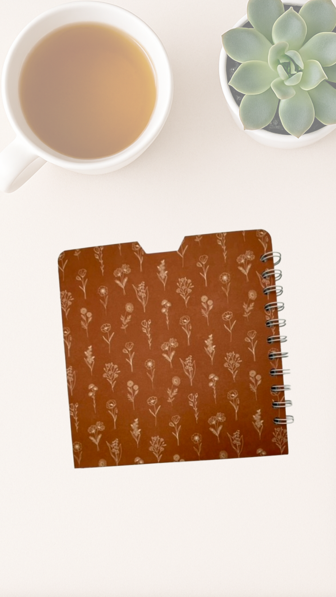 Notebook with back cover floral pattern next to a cup of tea and a succulent plant on a white surface