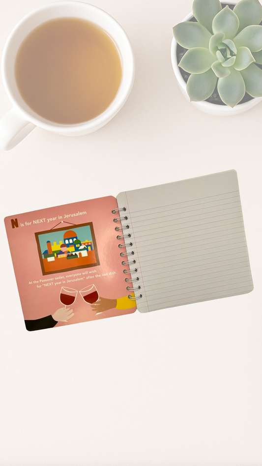 Notebook with a pink cover featuring a colorful illustration celebrating Jerusalem, next to a cup of tea and a succulent plant on a white surface.