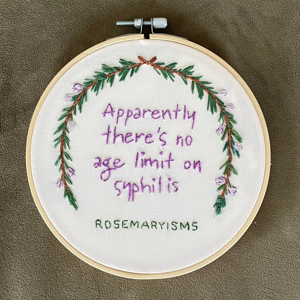 A circular embroidery hoop containing a white fabric background with purple text reading 'Apparently there's no age limit on syphilis' and a green floral wreath, mounted on a textured taupe fabric background. The brand name 'ROSEMARYSMS' is stitched at the bottom.