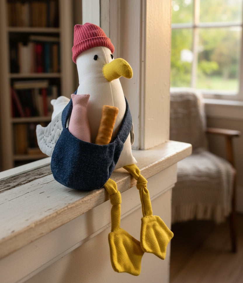 A plush seagull toy standing on a small wooden block against a bright blue backdrop. The plush has a white body, yellow beak and legs, gray wings, and wears a pink knit hat and a dark blue sling bag holding a pink plush item and a yellow stick.