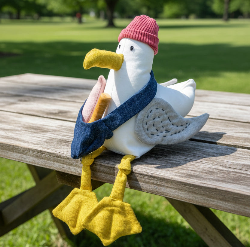 A plush stuffed seagull toy wearing a pink knit beanie, dark blue scarf, and yellow beak/legs, posed on a wooden block against a bright blue studio background.