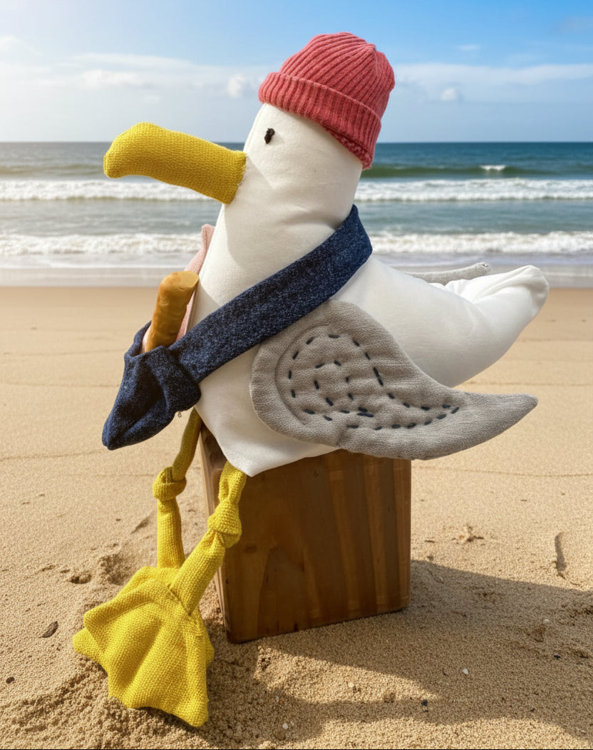 A plush toy seagull sitting on a wooden block against a bright blue studio background. The seagull is white with a yellow beak and webbed feet, wearing a pink knit hat and a dark blue scarf.