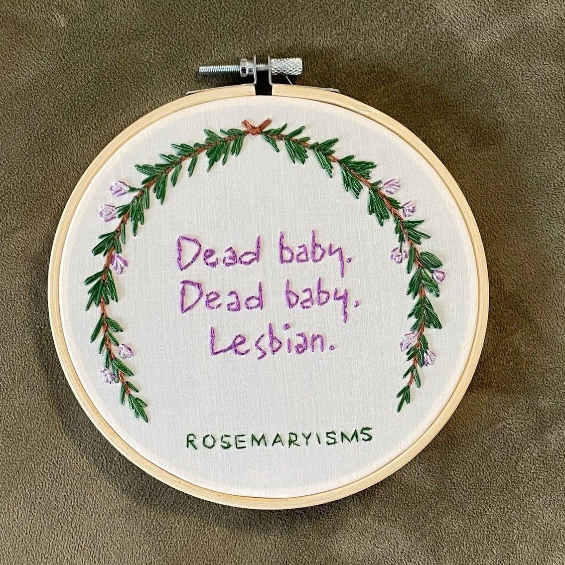 A circular embroidery hoop containing a white fabric with purple text reading 'Dead baby, dead baby, lesbian.' surrounded by a green leaf wreath and small purple flowers.