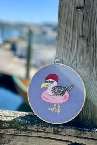Embroidery hoop with a embroidered seagull wearing a red hat on a wooden surface with a blurred waterfront background.