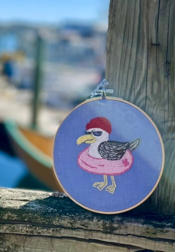 Embroidery of a bird wearing a red hat and pink dress in an embroidery hoop, set against a blurred waterfront background.