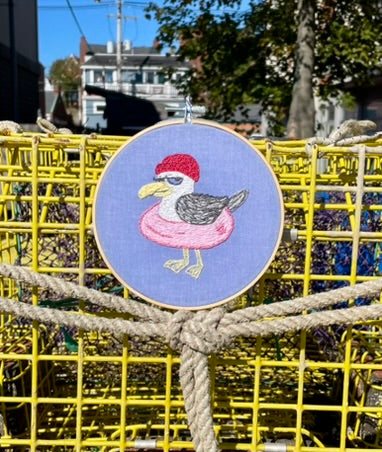 Embroidery hoop with a seagull design on a blue background, hanging on yellow lobster traps.
