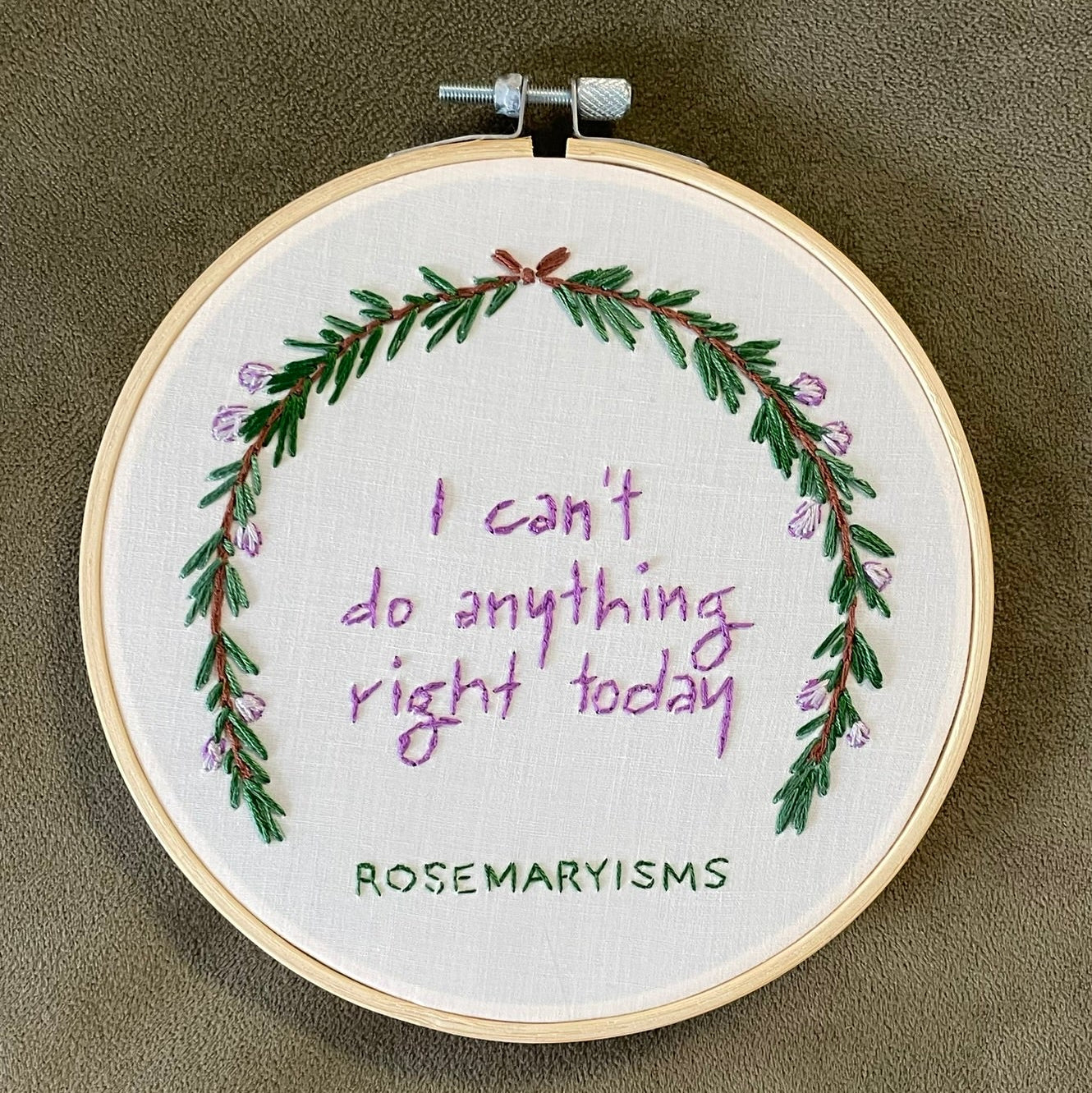 A circular embroidery hoop containing a white fabric with purple text reading 'I can't do anything right today' surrounded by a green wreath and small purple flowers. The hoop is placed on a textured olive-brown surface, with the brand name 'ROSEMARYS' printed vertically on the left side.