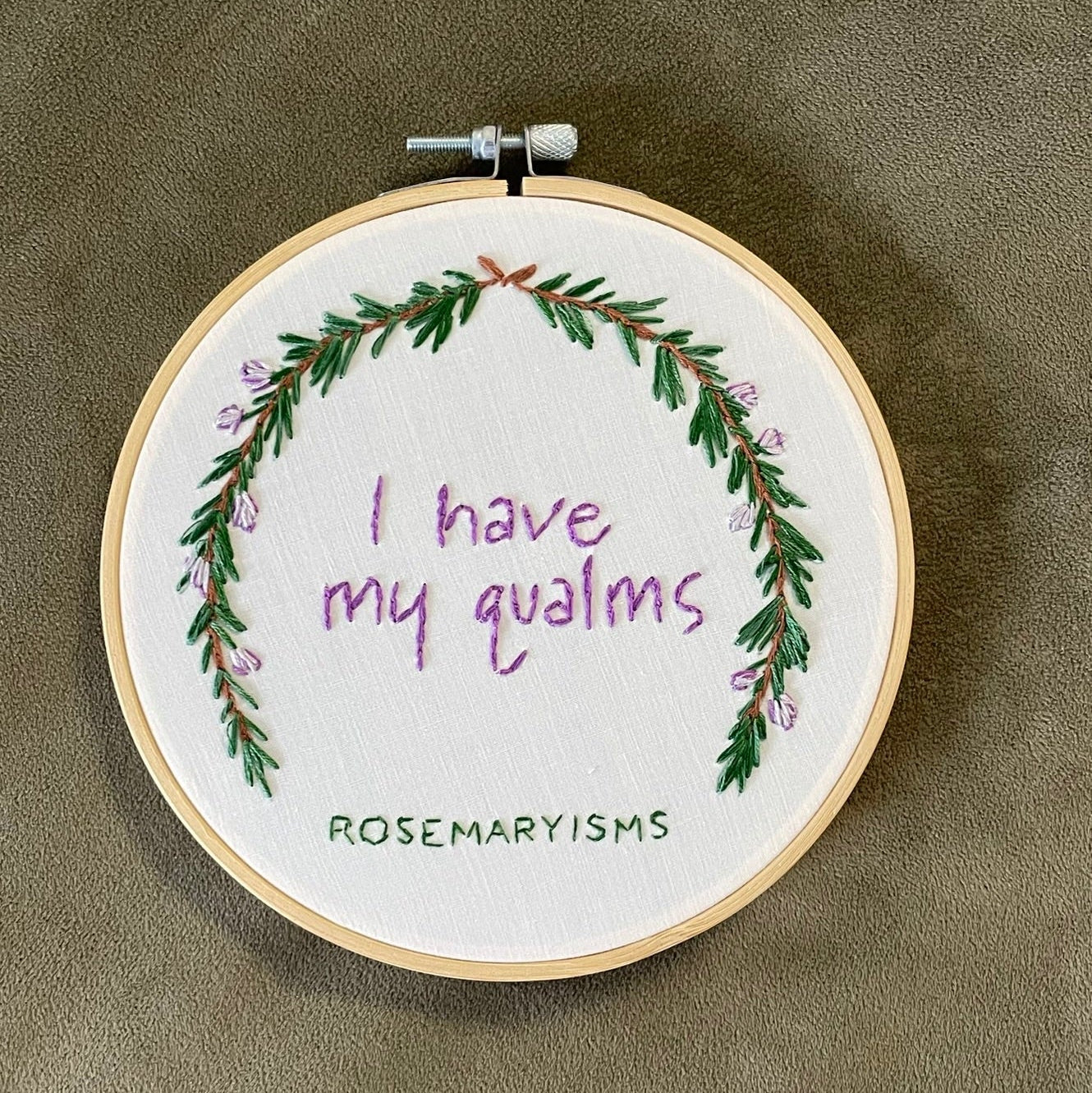 A circular embroidery hoop containing a white fabric with a green floral wreath and purple cursive text reading 'I have my qualms', shown on a textured taupe background. The brand name 'ROSEMARisms' is printed vertically on the left side of the hoop.
