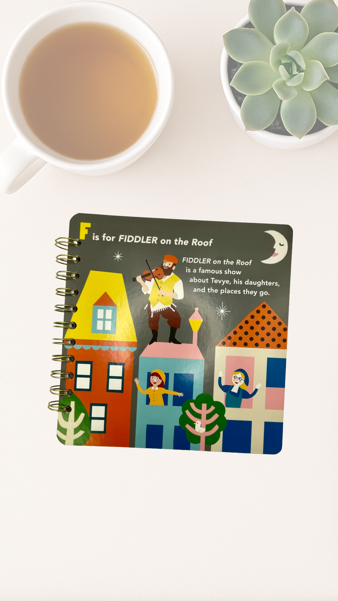 Notebook with 'F is for Fiddler on the Roof' cover design on a white surface with a cup of tea and succulent plant.