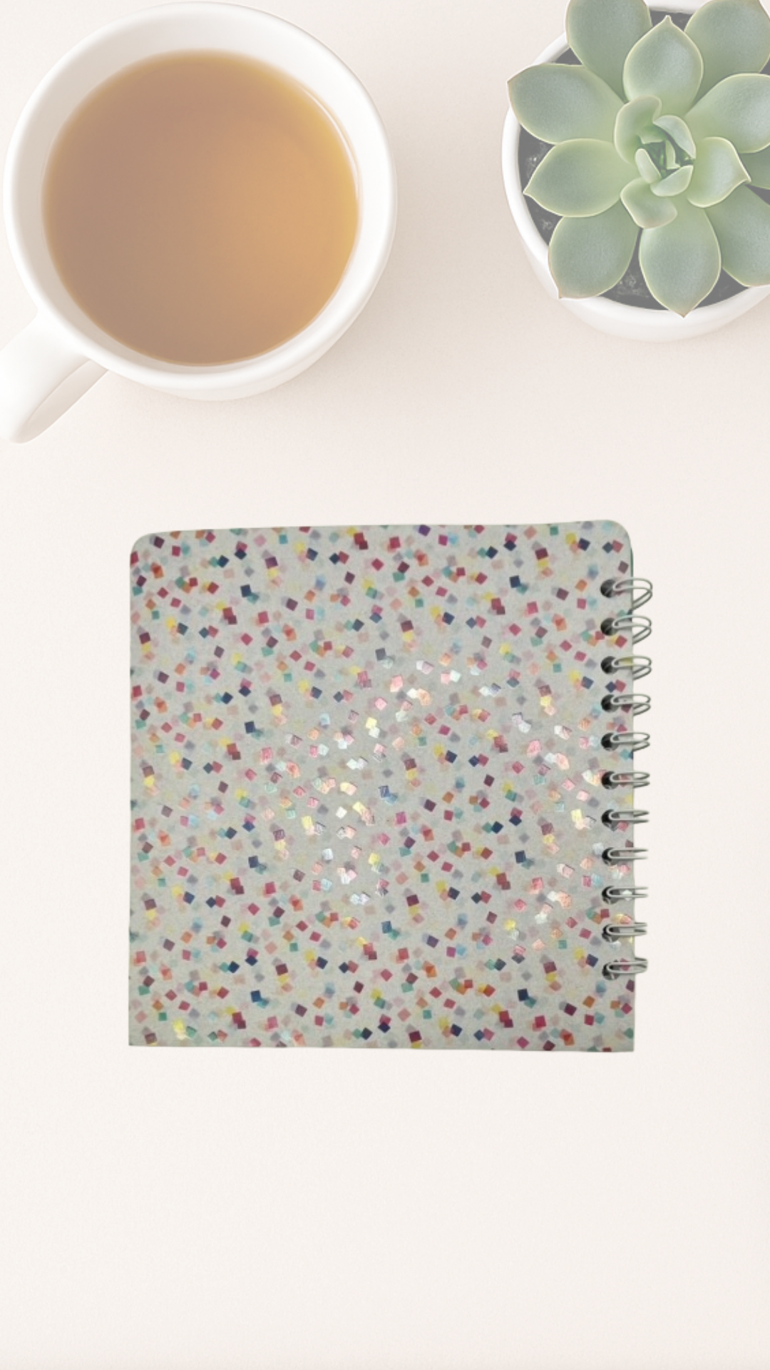 Notebook with colorful confetti pattern back cover next to a cup of tea and a succulent plant on a white surface