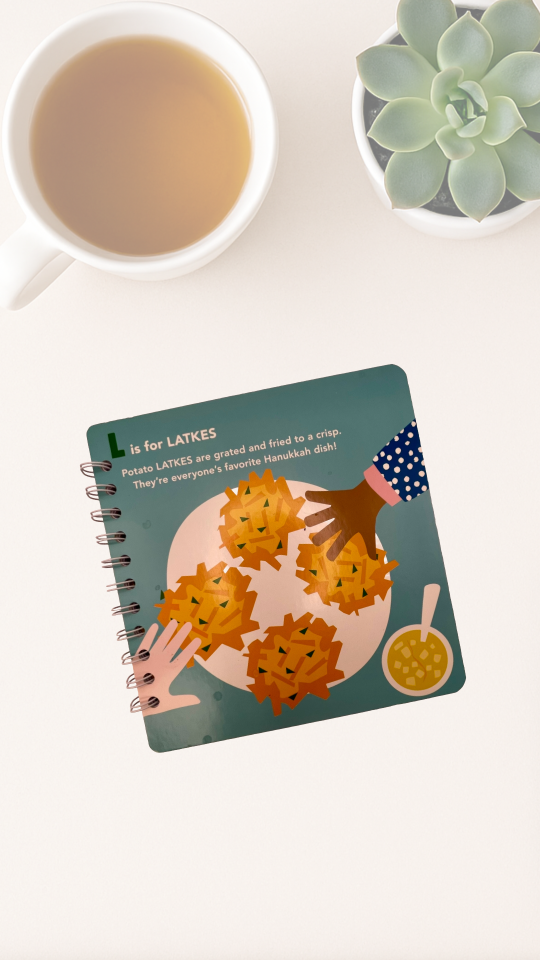 Notebook with potato latke illustration on a white surface with a cup of tea and plant.