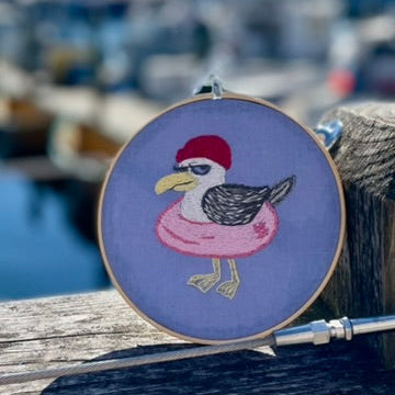 Embroidery hoop with a seagull design on a dock with boats in the background