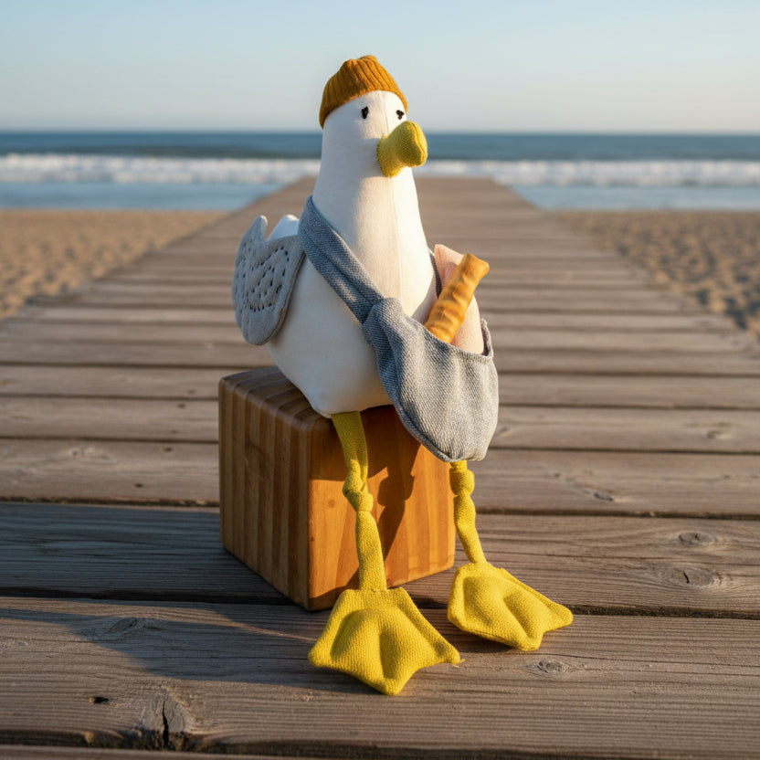 A plush seagull toy wearing a mustard yellow beanie, yellow webbed feet, and a gray crossbody bag, posed on a brown suitcase against a blue gradient background.