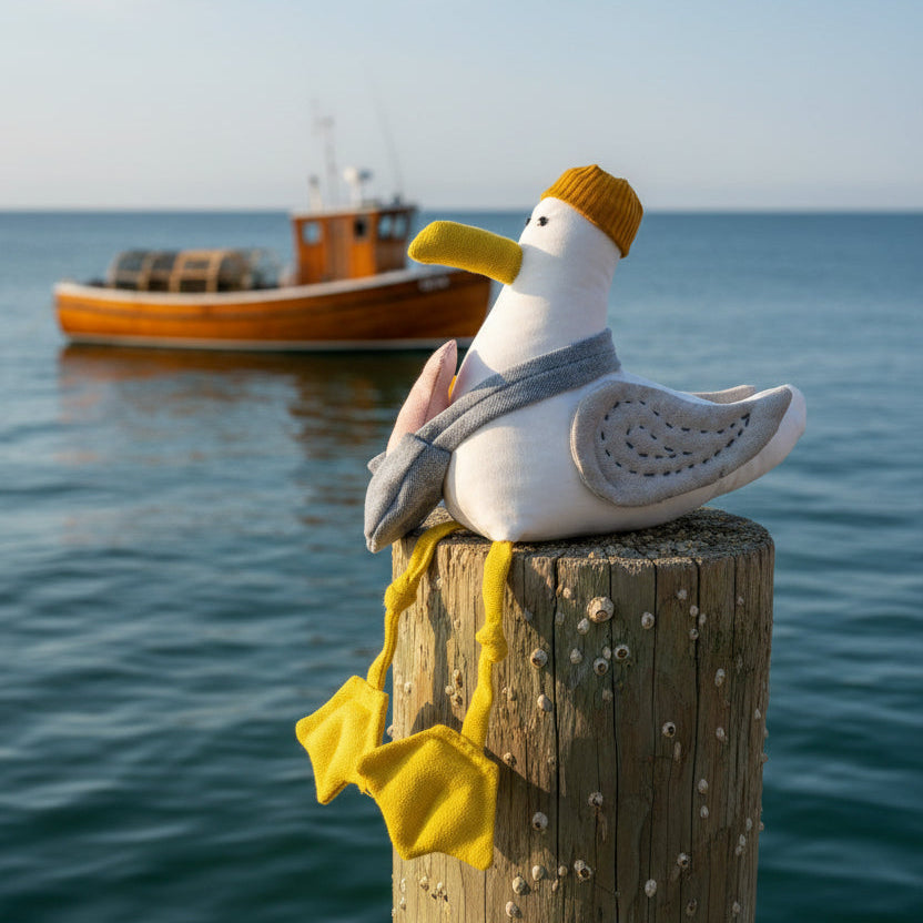 A plush seagull toy wearing a mustard yellow knit hat, gray scarf, and yellow webbed feet, posed on a small wooden block against a bright blue studio background.