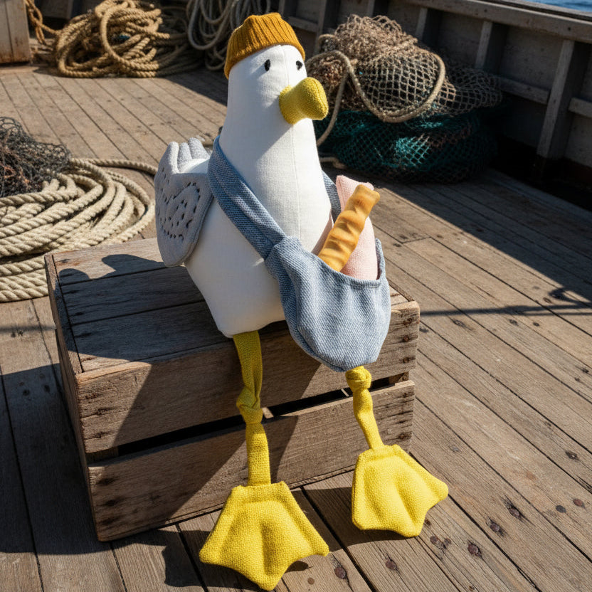 A plush stuffed bird toy with a white body, yellow beak and feet, wearing a mustard yellow knit hat and gray wrap, holding a pink bag, posed on a small brown box against a bright blue studio background.