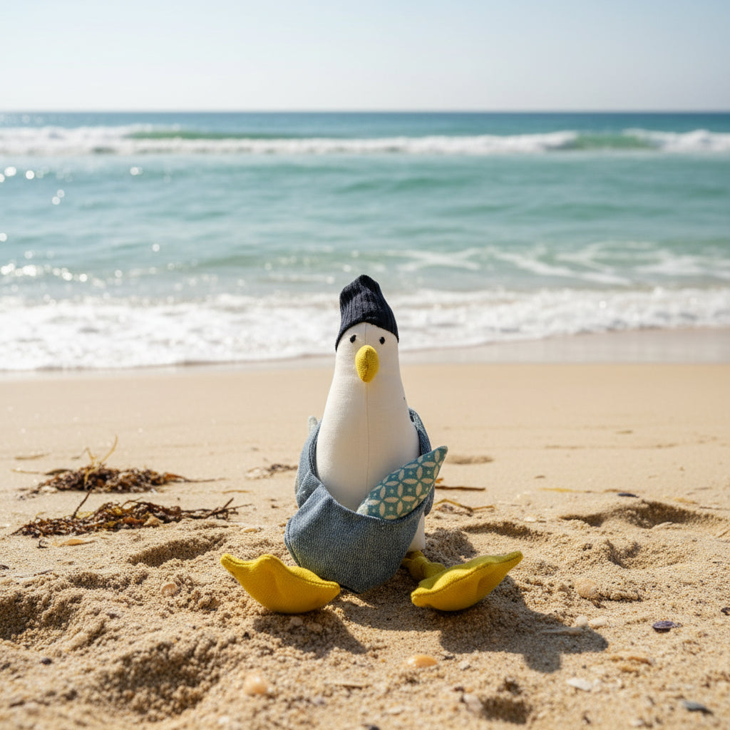 A handmade plush toy seagull wearing a navy beanie, light denim bag, and yellow webbed feet, sitting on a beach with a patterned fish