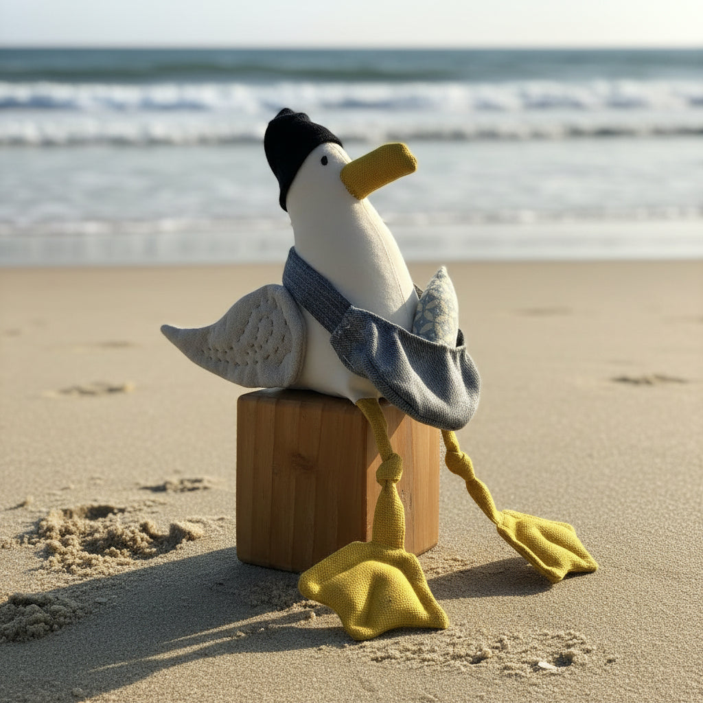 A handmade felt seagull plush toy wearing a navy beanie, light denim bag, and yellow webbed feet, sitting on a wooden block inside a blue lightbox studio setup.
