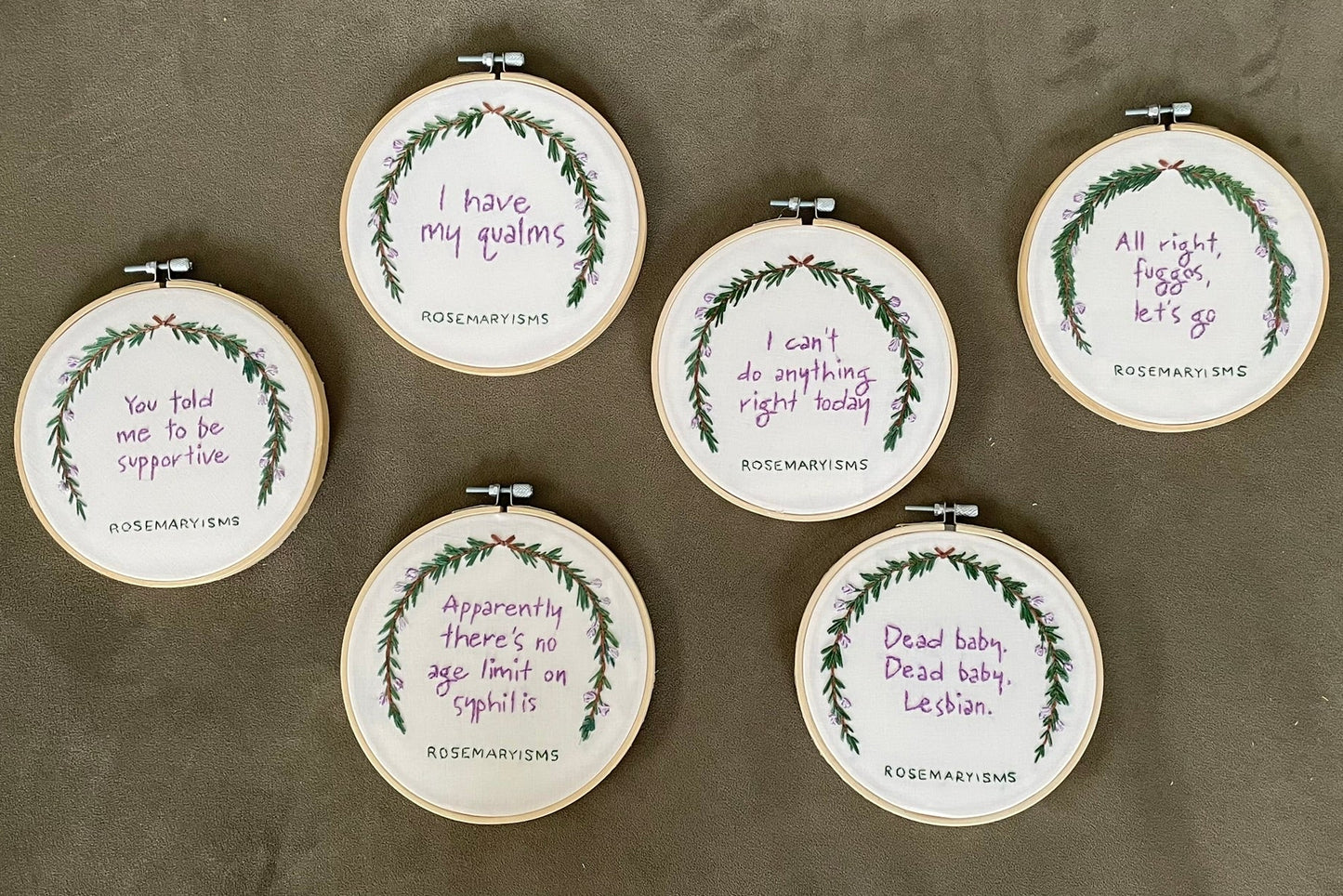 Six round embroidery hoops arranged on a textured gray surface, each containing a white fabric background with a green wreath design and purple handwritten text.