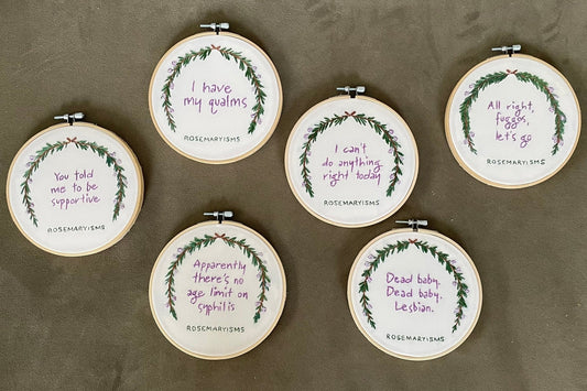 Six round embroidery hoops arranged on a textured gray surface, each containing a white fabric background with a green wreath design and purple handwritten text.
