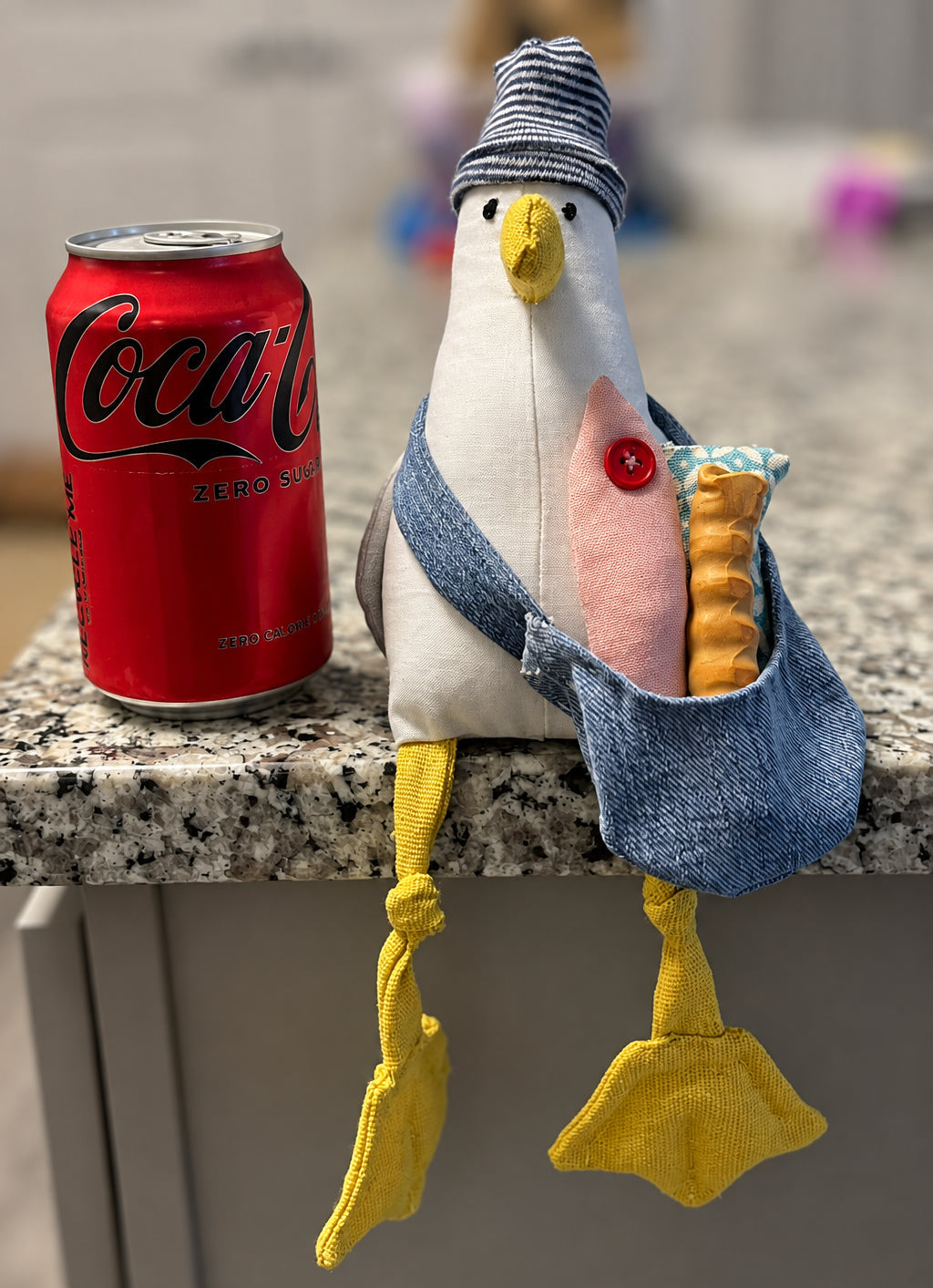 A handmade plush toy of a white seagull wearing a blue denim bag and striped hat, sitting on a granite kitchen counter next to a red Coca-Cola can.