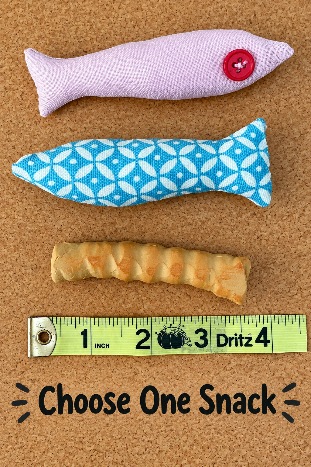 Three small handmade fabric fish-shaped toys in different colors and patterns are laid out on a brown cork background, with a yellow measuring tape below them for scale.