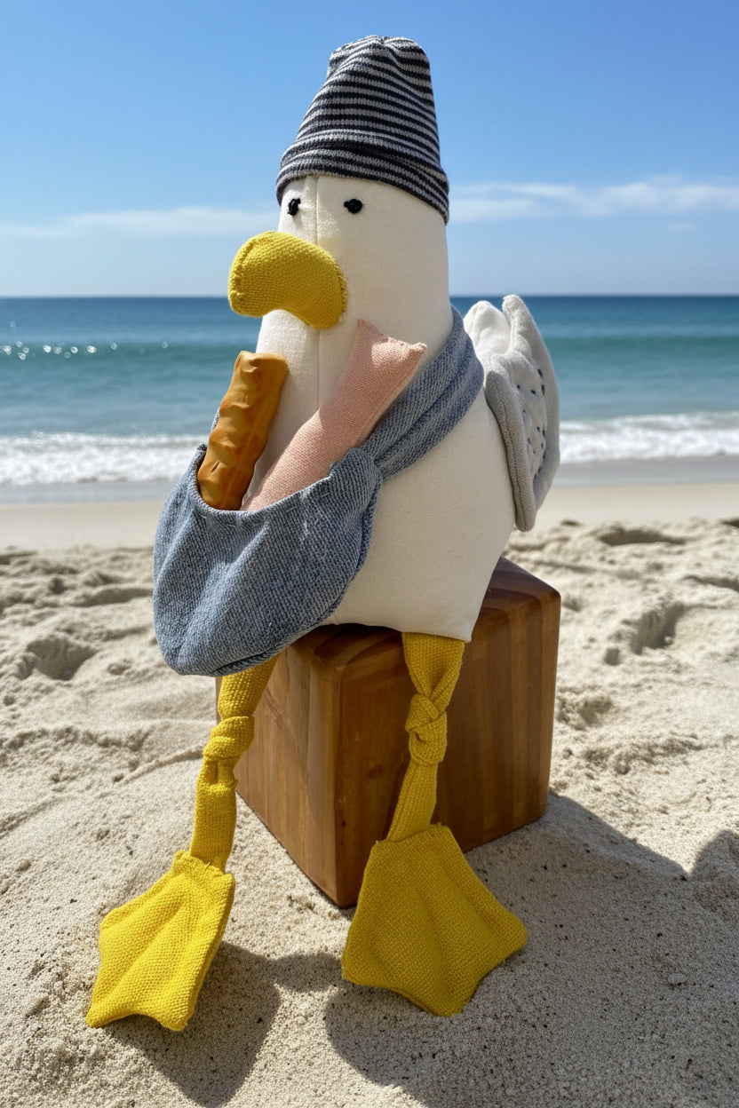 A plush toy seagull sitting on a small wooden box against a bright blue studio background. The toy has a white body, yellow beak and feet, a gray sleeve, and a striped gray-and-white hat.