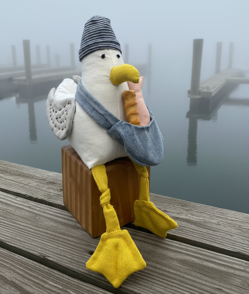 A plush stuffed bird toy sitting on a wooden block against a bright blue studio background. The bird has a white body, yellow beak and feet, a gray scarf, a striped gray and white hat, and a pink-and-orange striped stuffed hand held to its beak.