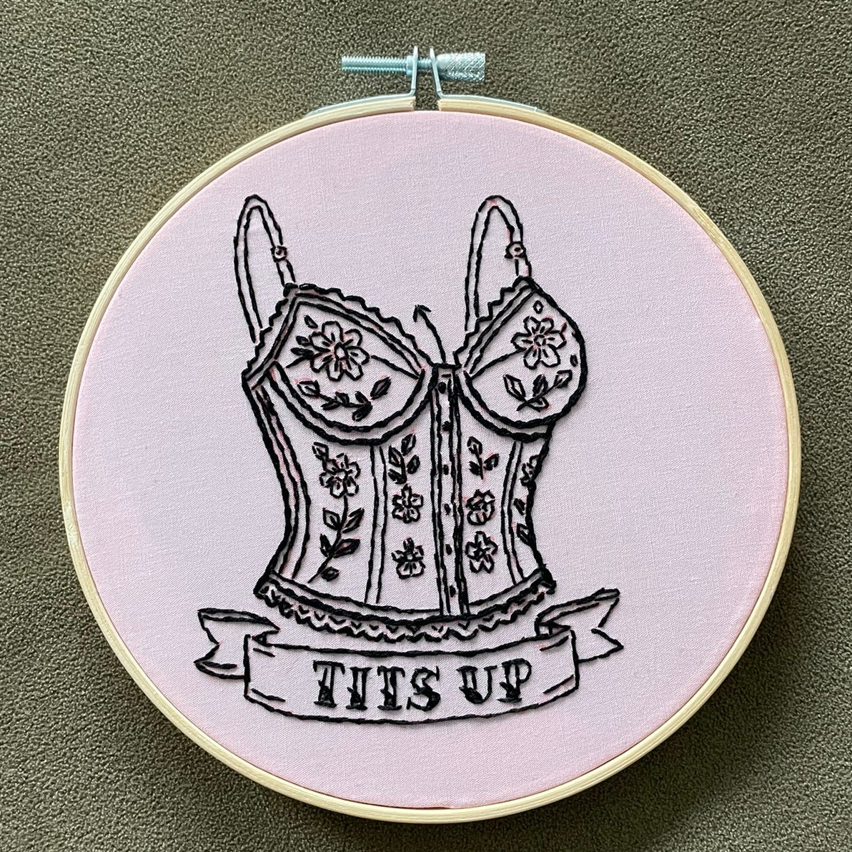 Embroidery hoop with a black and white illustration of a corset and 'TITS UP' text on a green background