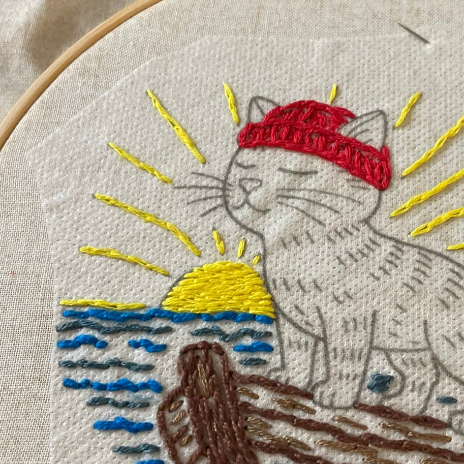 Embroidery of a cat on a log with a red hat in an embroidery hoop.
