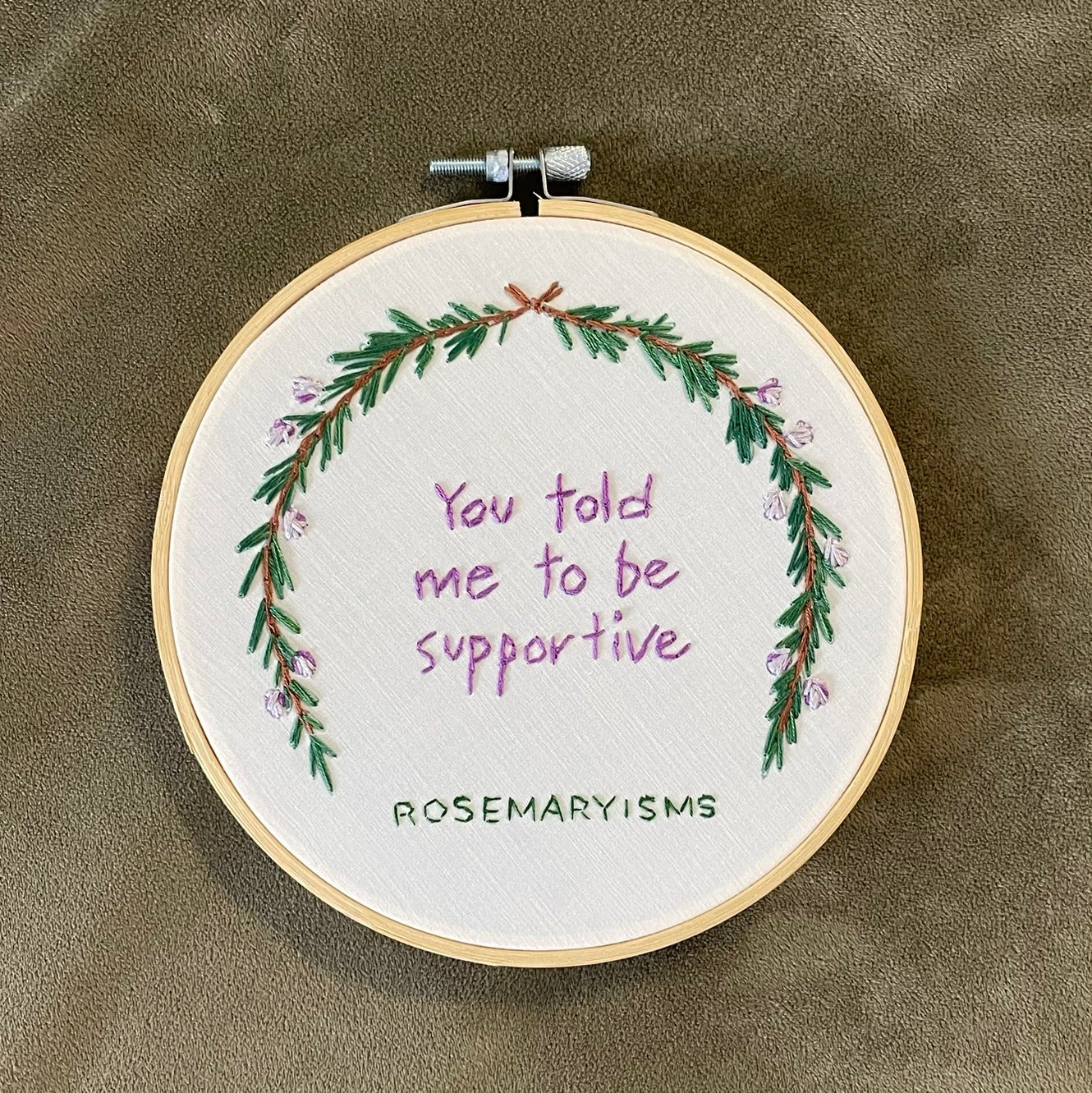 A circular embroidery hoop containing a white fabric with a green floral wreath and purple text reading 'You told me to be supportive', photographed from above on a textured olive-brown surface. The hoop has a wooden frame, a white center, and a metal clamp on the right side.